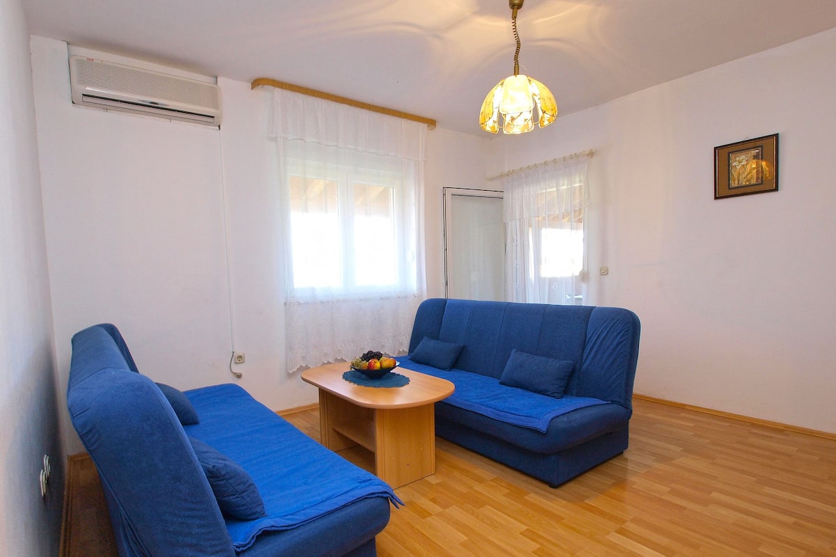 A room with a blue sofa, a smaller blue chair, a wooden coffee table with fruit, and a hanging light fixture.