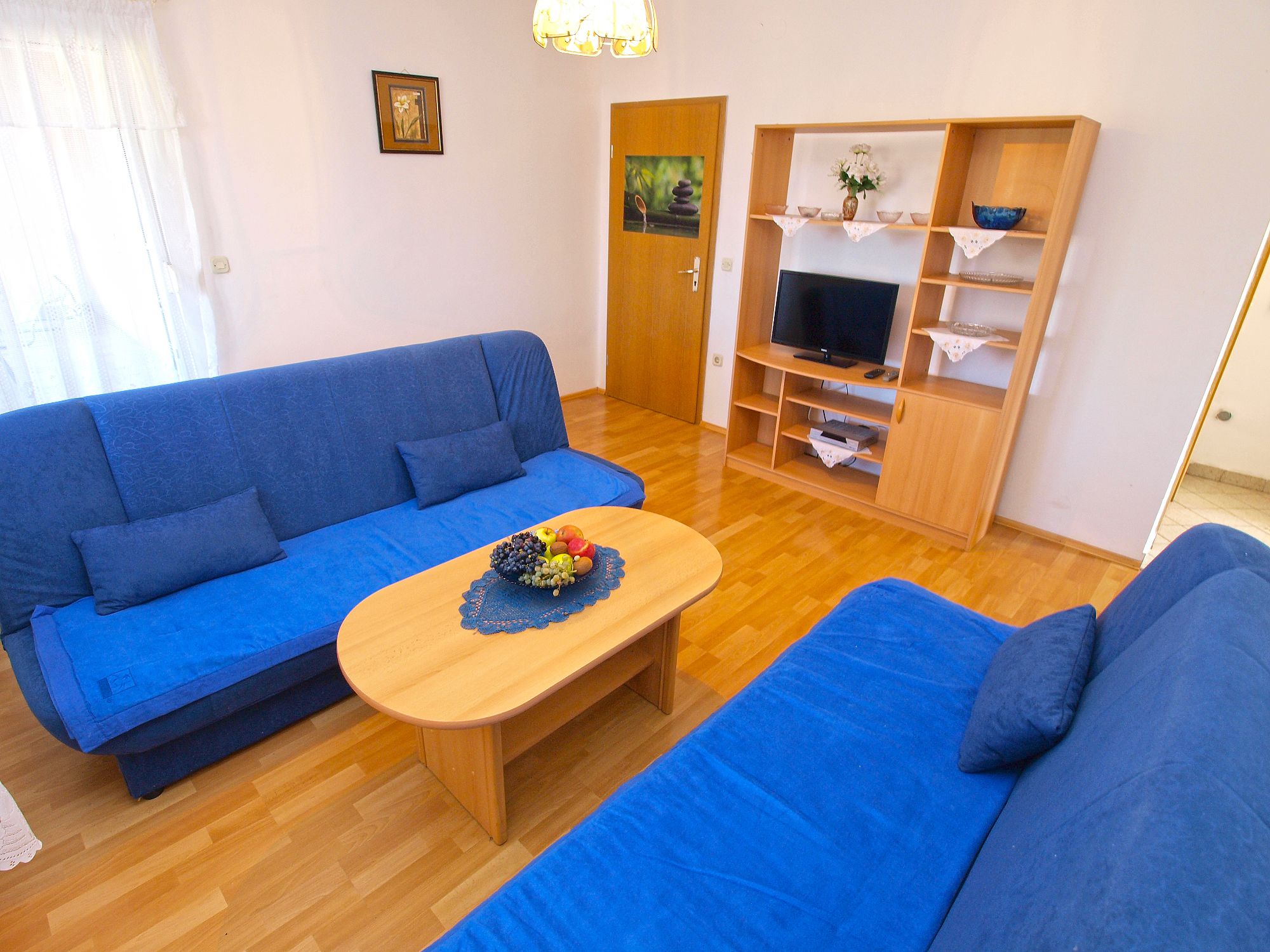A living room with a blue sofa set, a wooden coffee table with fruit, a TV stand with shelves, and a door with a painting.
