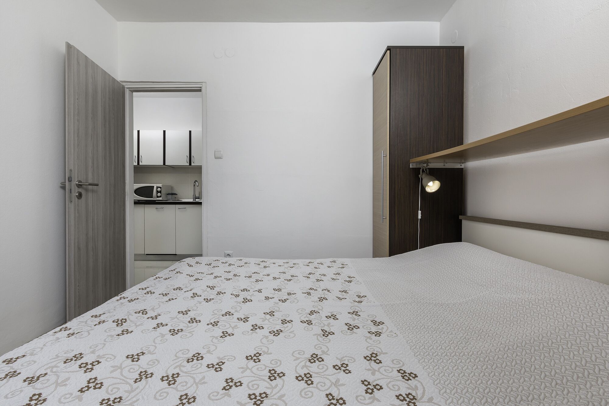 A bedroom with a bed, a nightstand, a wardrobe, and a kitchenette in the background.