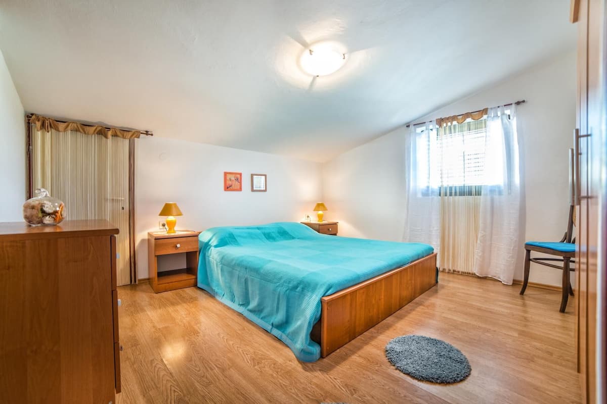 A bedroom with a wooden bed, two bedside tables with lamps, a wooden wardrobe, a small rug, and a window with curtains.