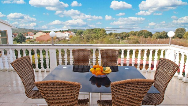 A balcony with wicker chairs and a table, a bowl of fruit, and a scenic view of a coastal area with houses and trees. - Apartments Ivan 65 (Pula)