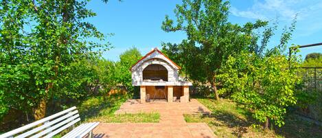 A stone-built outdoor oven with a tiled roof, surrounded by lush greenery and a paved path leading to it.
