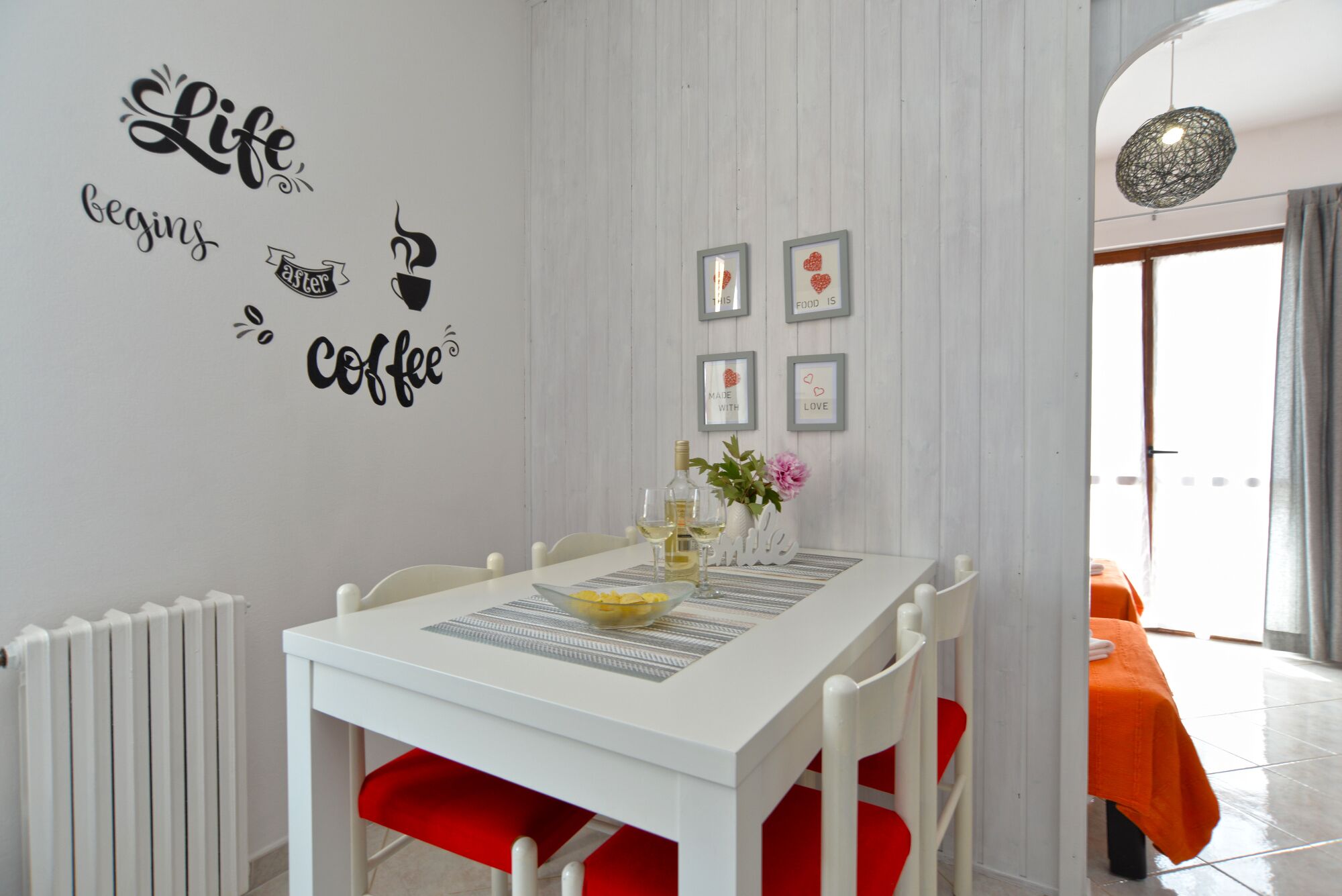 Un comedor con una mesa blanca, sillas rojas y un radiador blanco. Hay decoraciones en las paredes, incluyendo una frase y cuadros enmarcados.