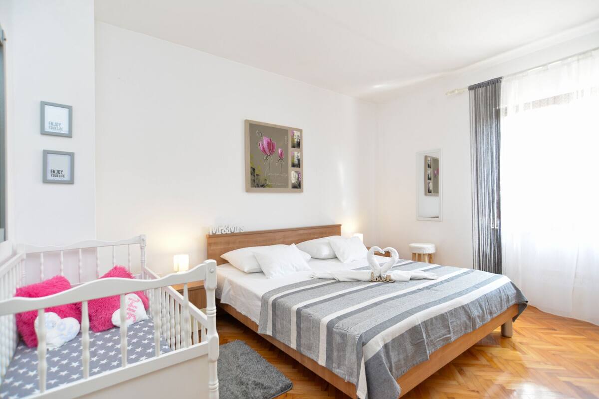 Ein Schlafzimmer mit einem Bett, einem Babybett, einem Nachttisch und einem Wandbild.