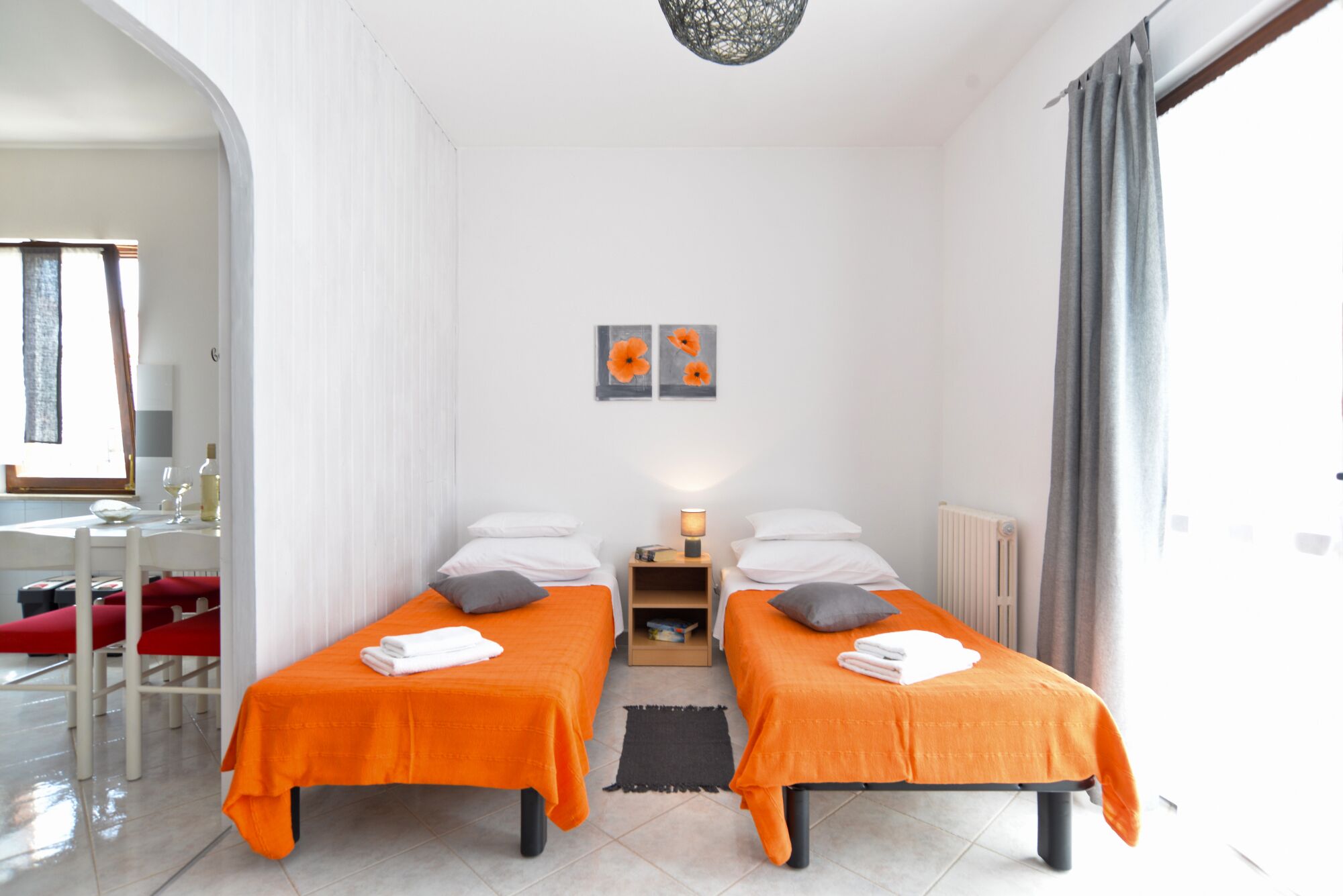 A hotel room with two beds, a small table, a chair, a radiator, and a window with curtains.