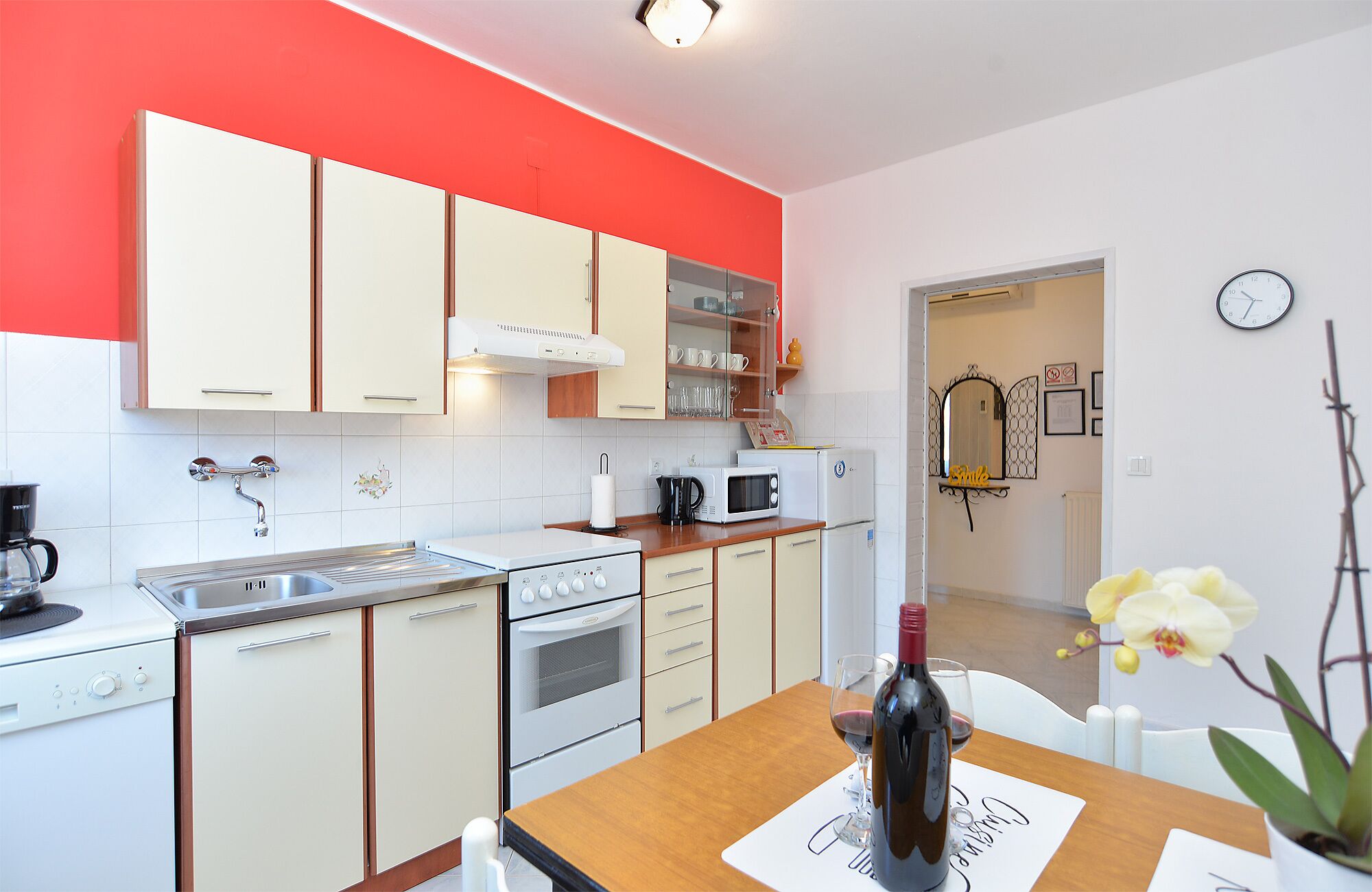 Una cocina moderna con armarios blancos y rojos, un microondas, un horno y un lavabo. Hay una mesa de comedor con una botella de vino y copas.