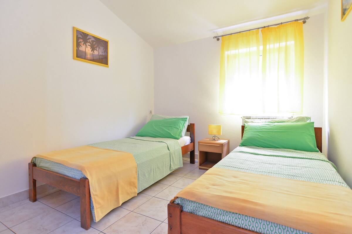 Una habitación con dos camas individuales, cada una con estructura de madera y ropa de cama verde. Hay una mesita de noche con una lámpara, una ventana con cortinas amarillas y un cuadro en la pared.