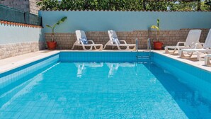 Una piscina con agua azul mosaico, rodeada de sillas de descanso blancas con cojines azules y plantas en macetas.