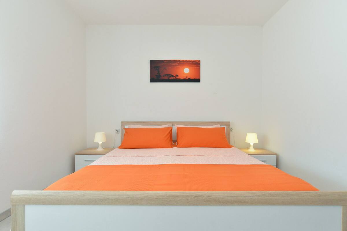 Un dormitorio con una cama, dos mesitas de noche con lámparas y un cuadro colgado en la pared que representa un atardecer.