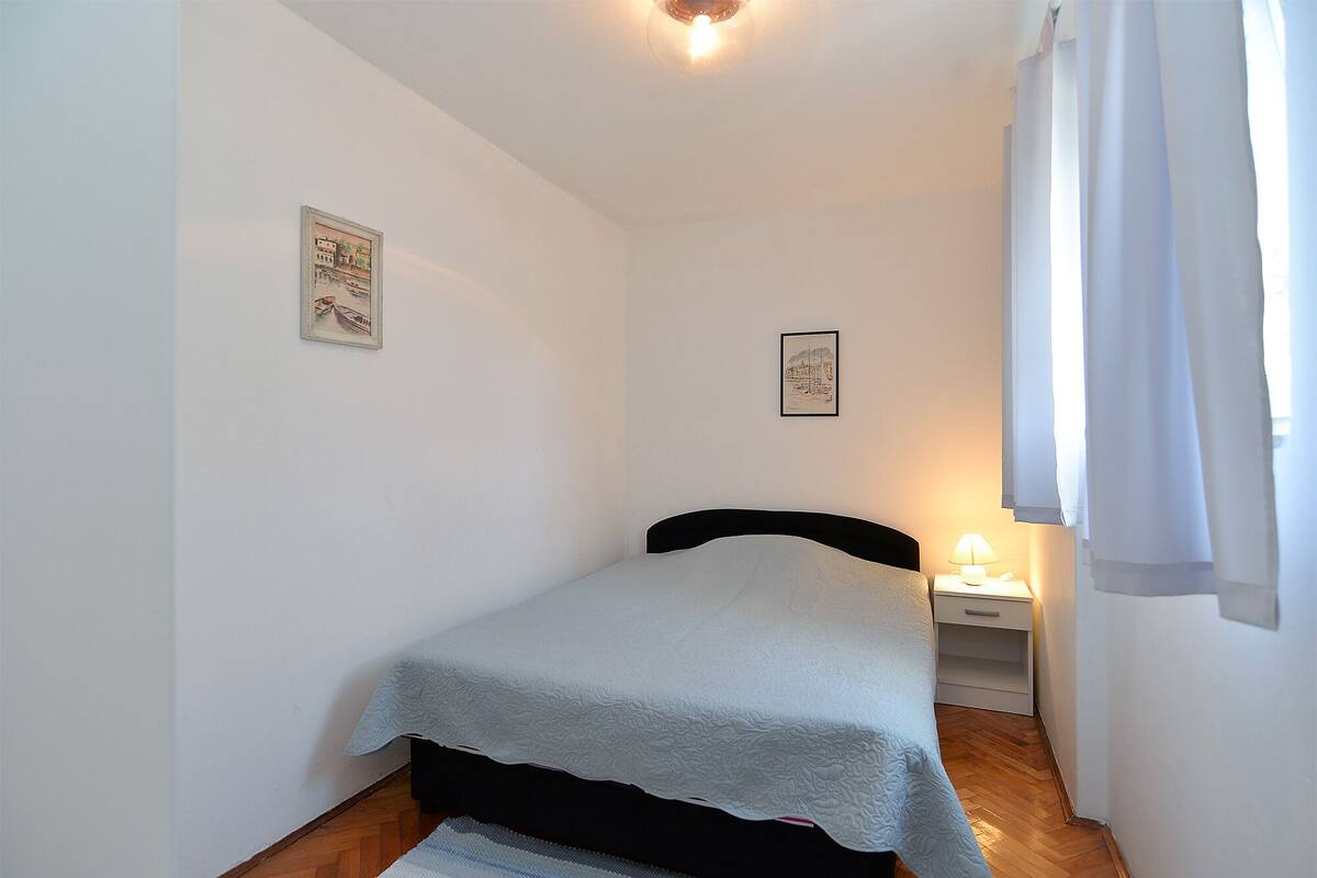 A small, single-bed room with a wooden floor, white walls, and two framed pictures. There is a bedside table with a lamp and a small drawer.