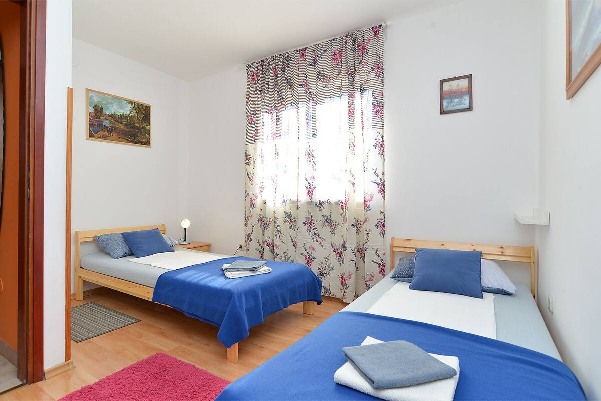A room with two beds, a wooden door, a painting, and a window with floral curtains.