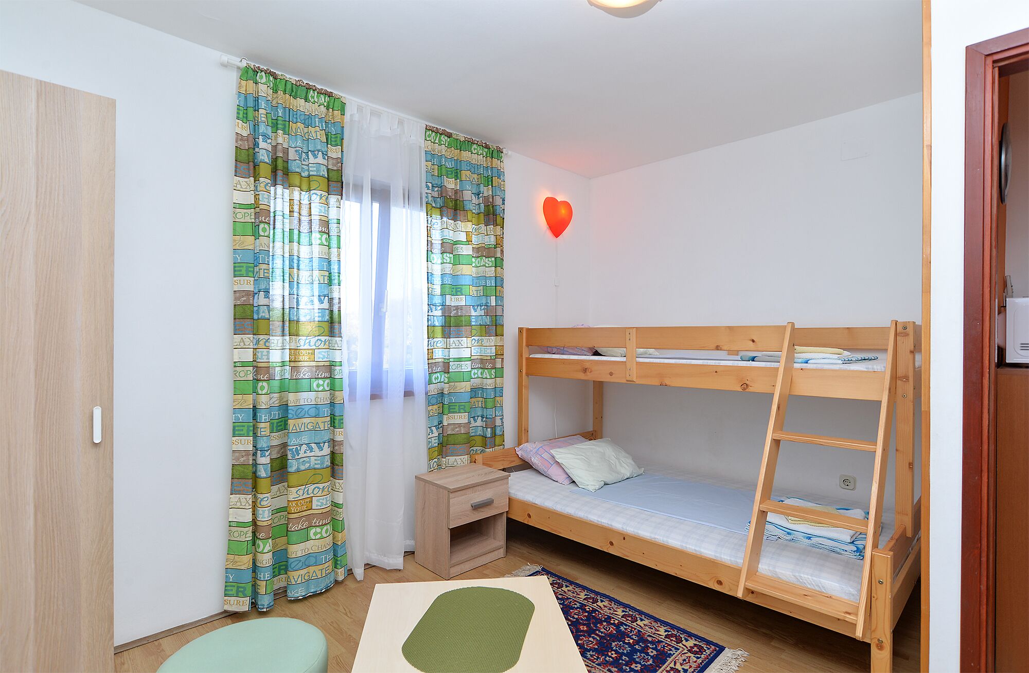 Una habitación con una cama individual superpuesta, una pequeña mesita de madera, un otoman verde y una cortina estampada.