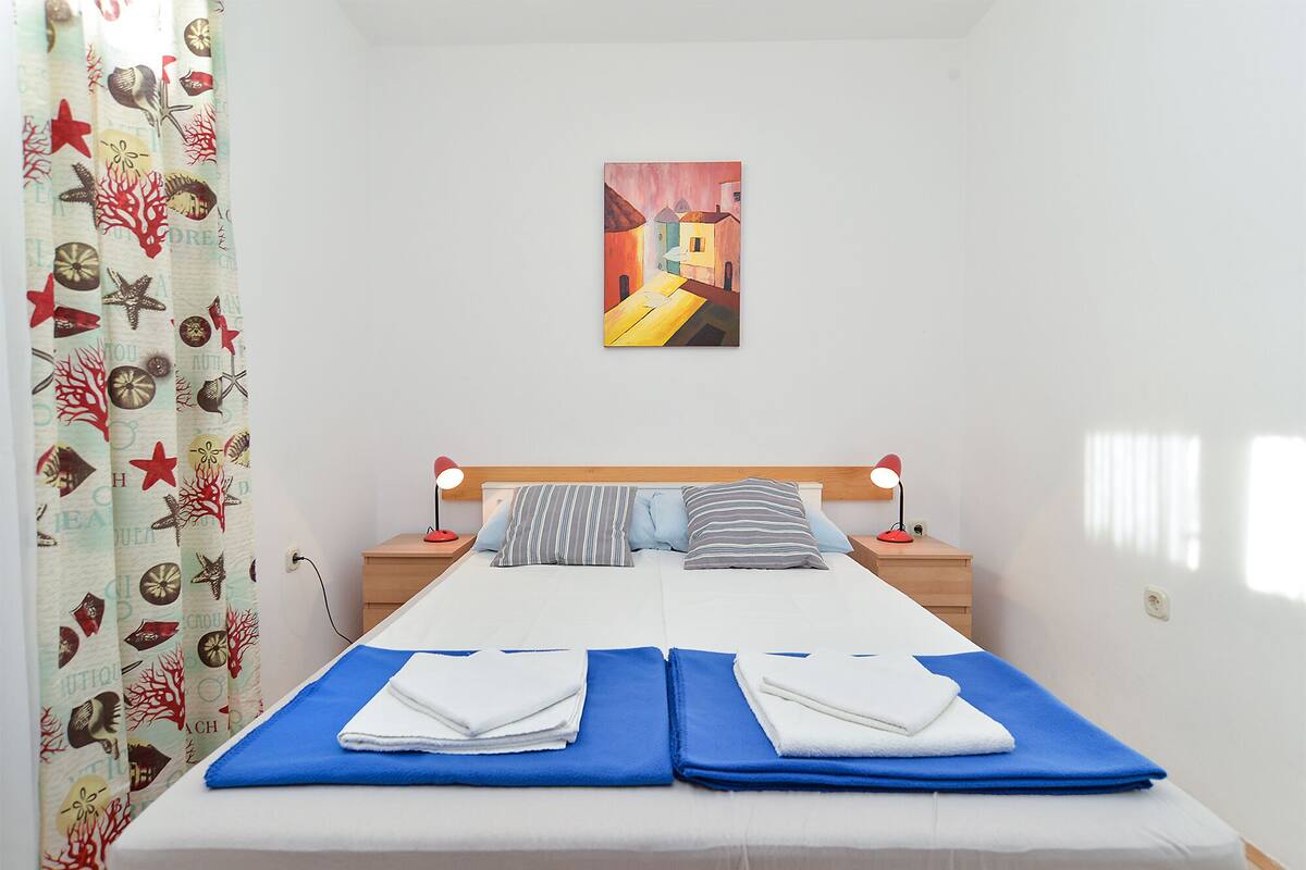 A bedroom with a bed, two bedside tables, a painting on the wall, and a curtain with a pattern.