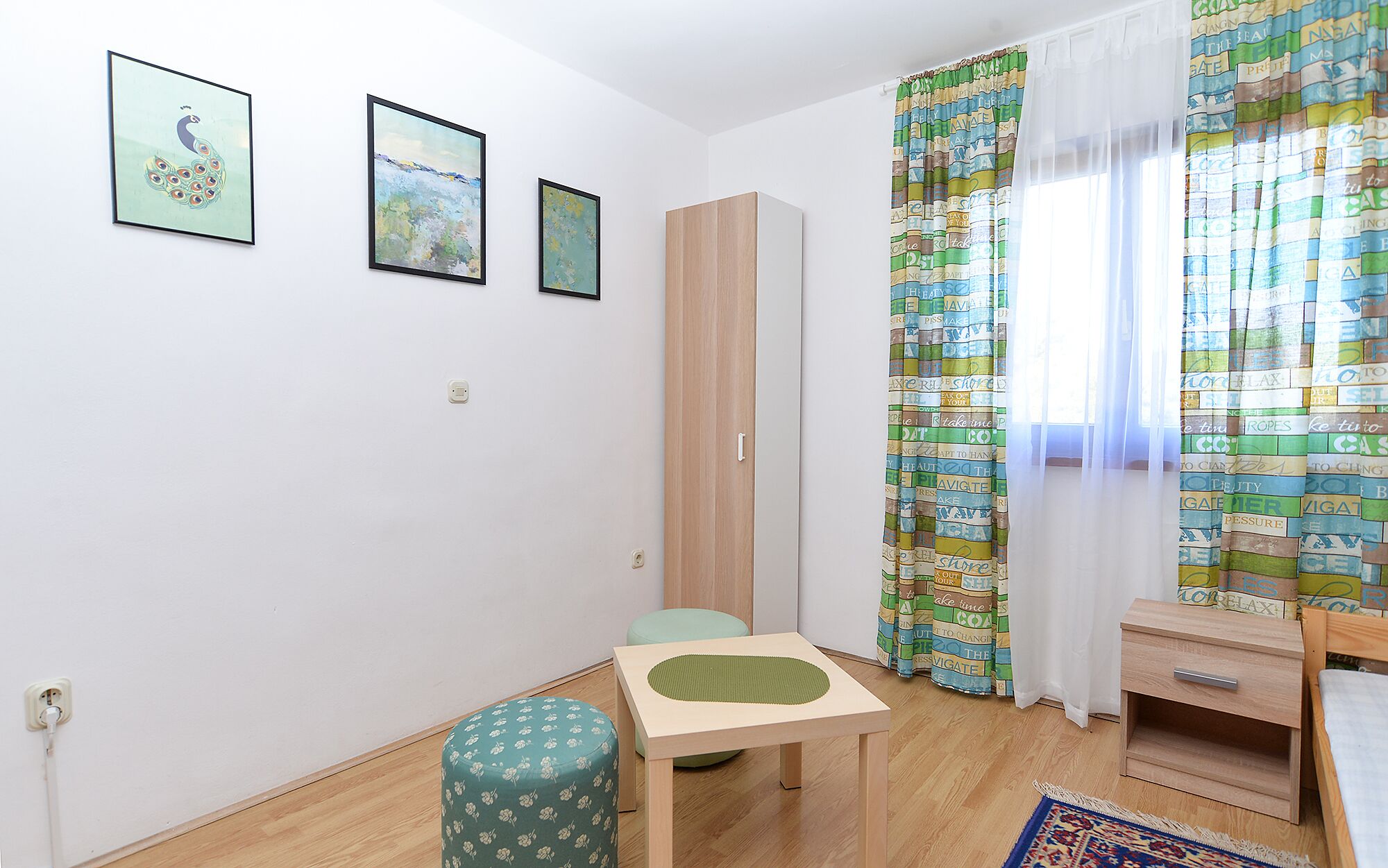 A small, tidy room with a wooden floor, a green patterned ottoman, a small table, a wardrobe, and a colorful curtain.