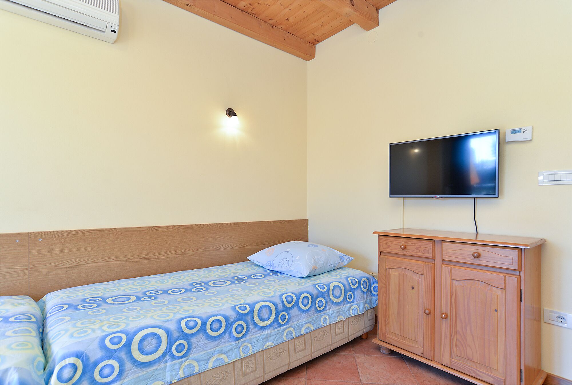 A single bed with a patterned bedspread, a wooden headboard, a television mounted on the wall, and a wooden cabinet.