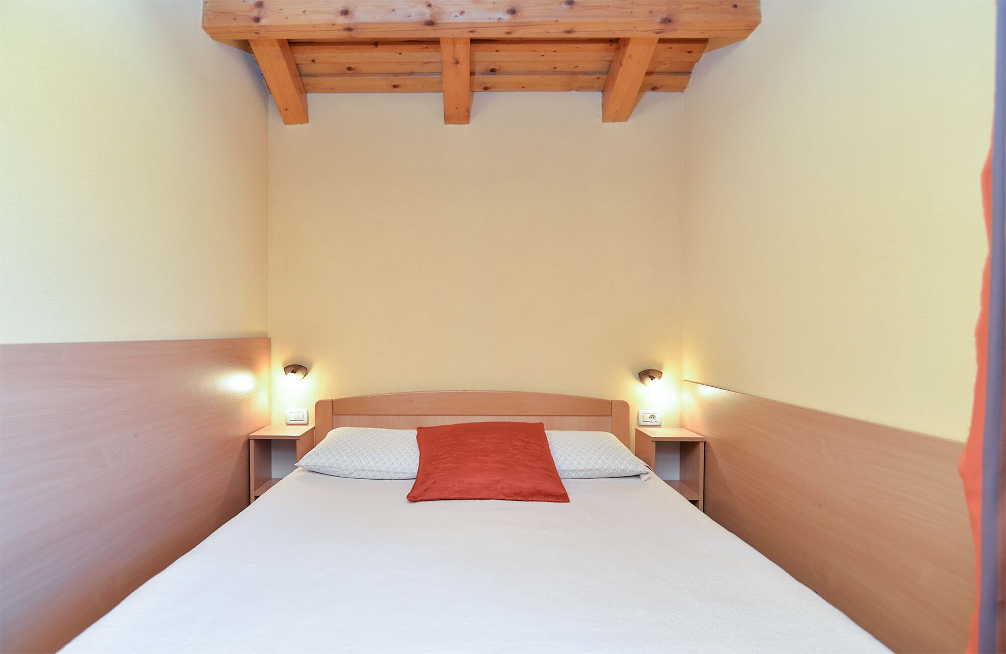A narrow room with a bed, wooden headboard, two bedside tables with lamps, and a wooden ceiling structure.