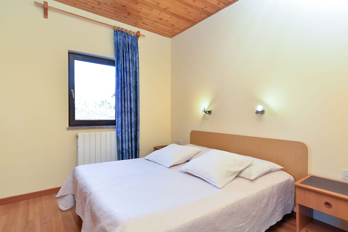 A bedroom with a wooden headboard, a window with curtains, a bedside table, and a wall-mounted light fixture.