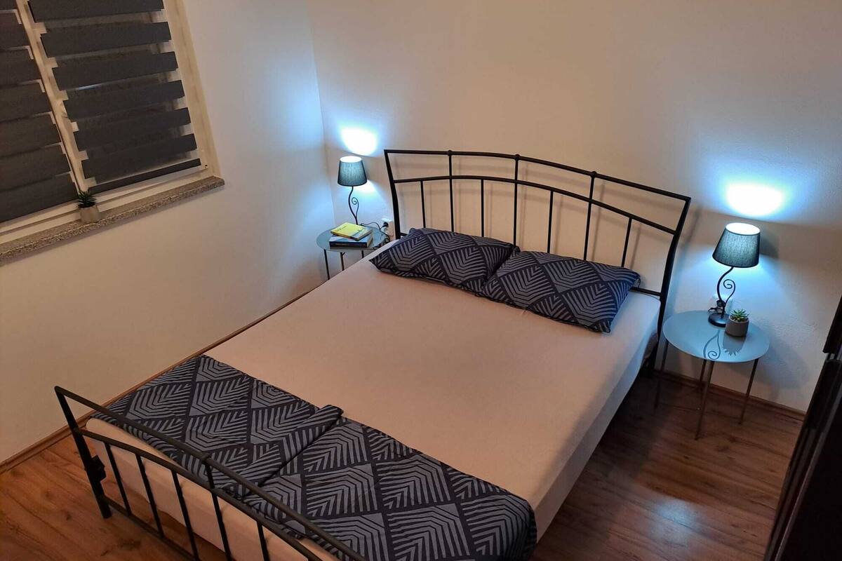 Un dormitorio pequeño y ordenado con una cama, dos mesitas de noche y una ventana con persianas.