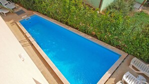 Una piscina rectangular con borde de azulejos, rodeada de sillas reclinables blancas y un seto bajo.