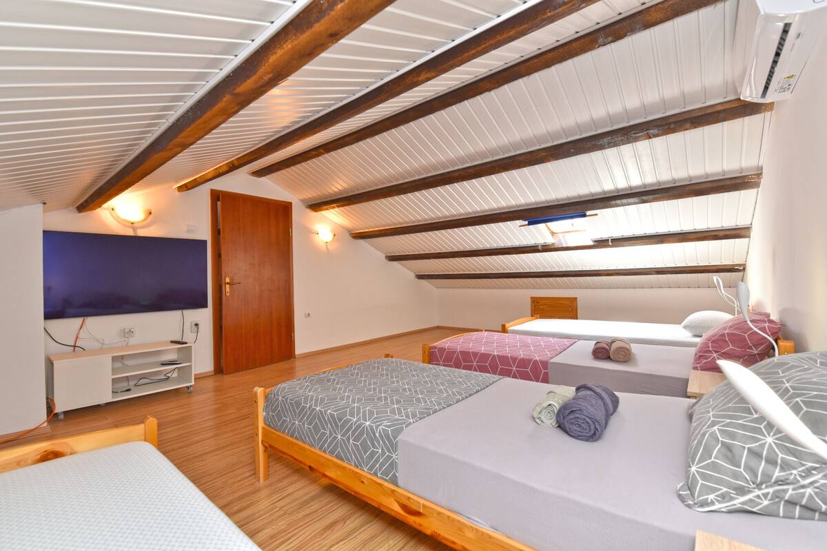 Un dormitorio con techo inclinado, vigas de madera, televisor, cama y mesita de noche.