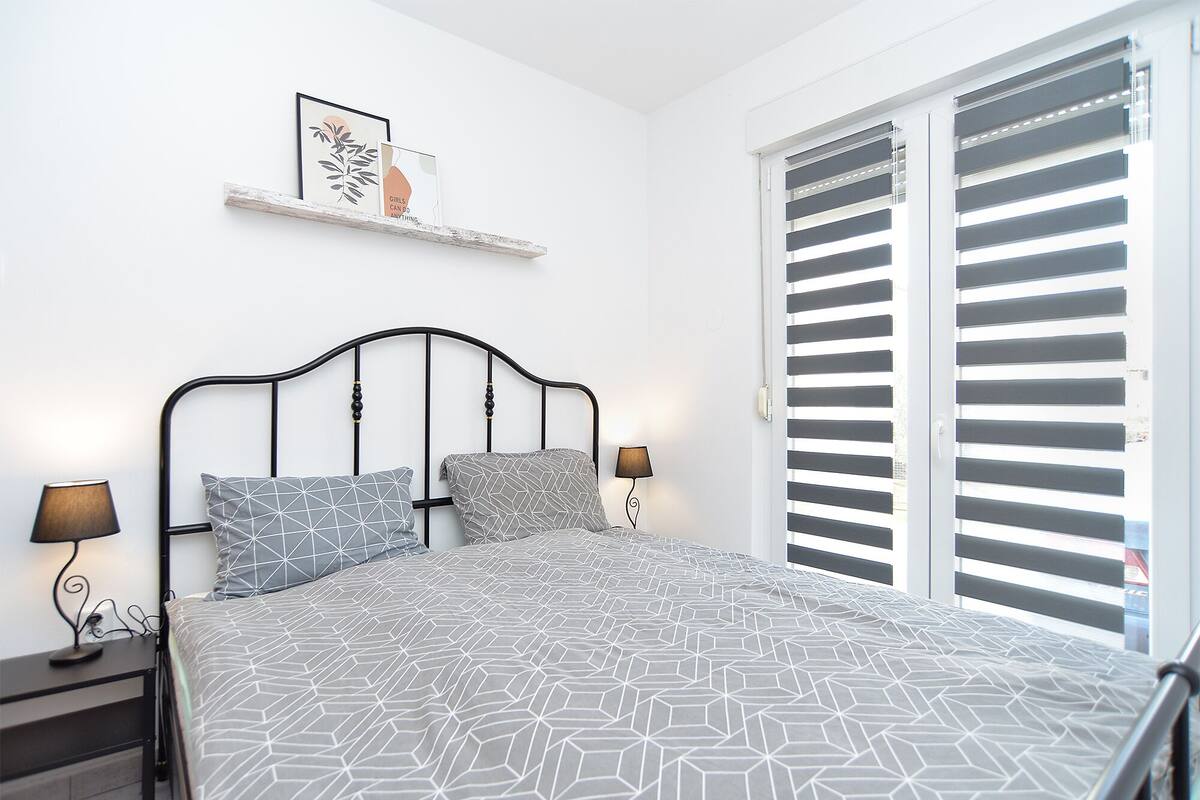 Un dormitorio con cama, dos lámparas de noche, un estante en la pared con obras de arte enmarcadas y una ventana con persianas.