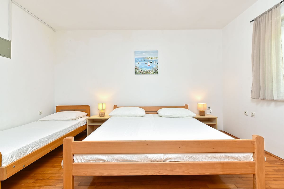 A room with two single beds, a wooden headboard, bedside tables with lamps, a small painting on the wall, and a window with curtains.