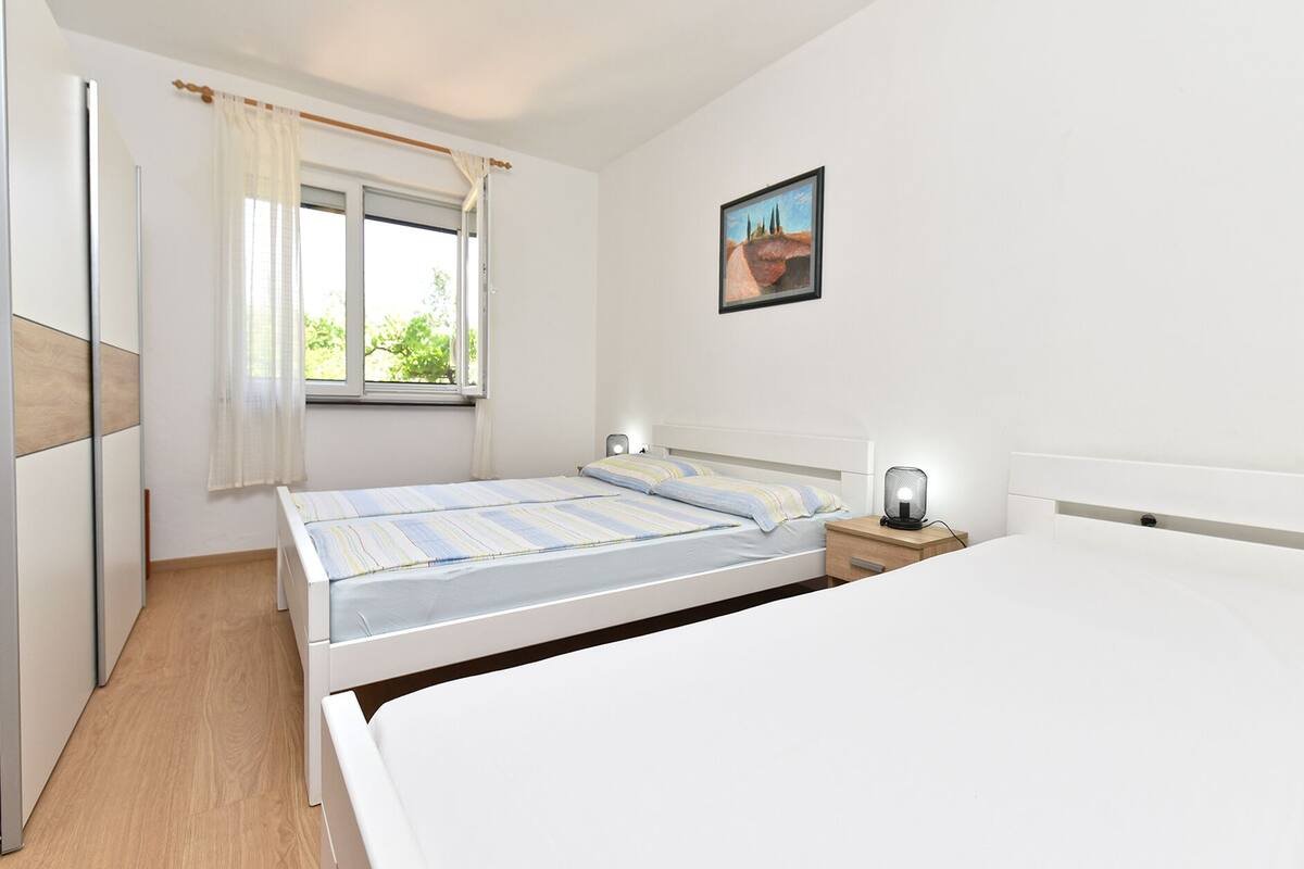 A bedroom with a large bed, a smaller bed, a bedside table with a lamp, a window with curtains, and a painting on the wall.