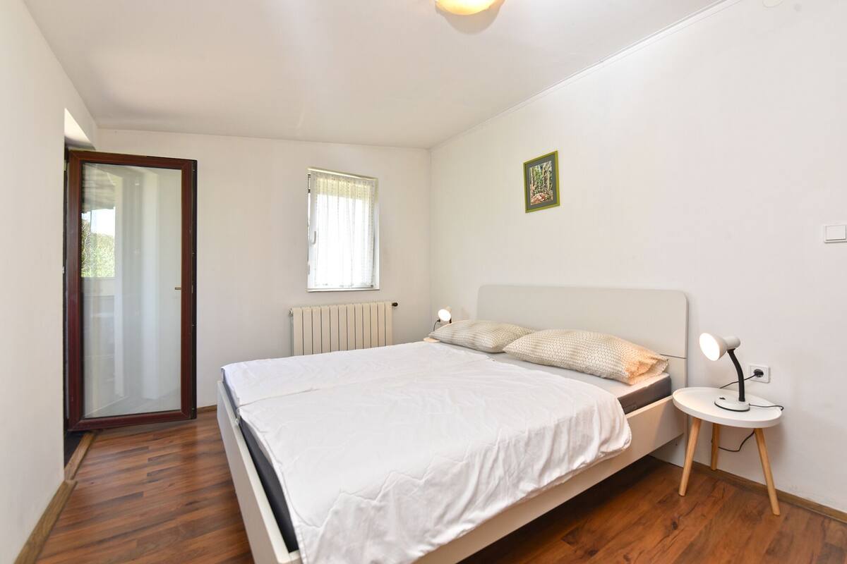 A bedroom with a bed, a bedside table, a lamp, a radiator, a window with curtains, and a door.