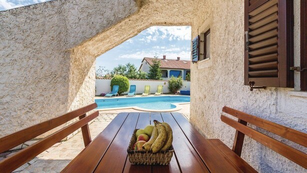 Una mesa de madera con una cesta de fruta en el centro, una piscina al fondo y vista de una casa a través de un arco.