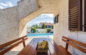 Taula de fusta amb una cistella de fruita al centre, una piscina de fons i vista d'una casa a través d'una llenca.