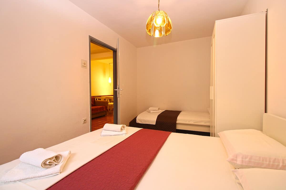 A hotel room with two beds, a red bed runner, white linens, a hanging light fixture, and a door leading to a dining area.