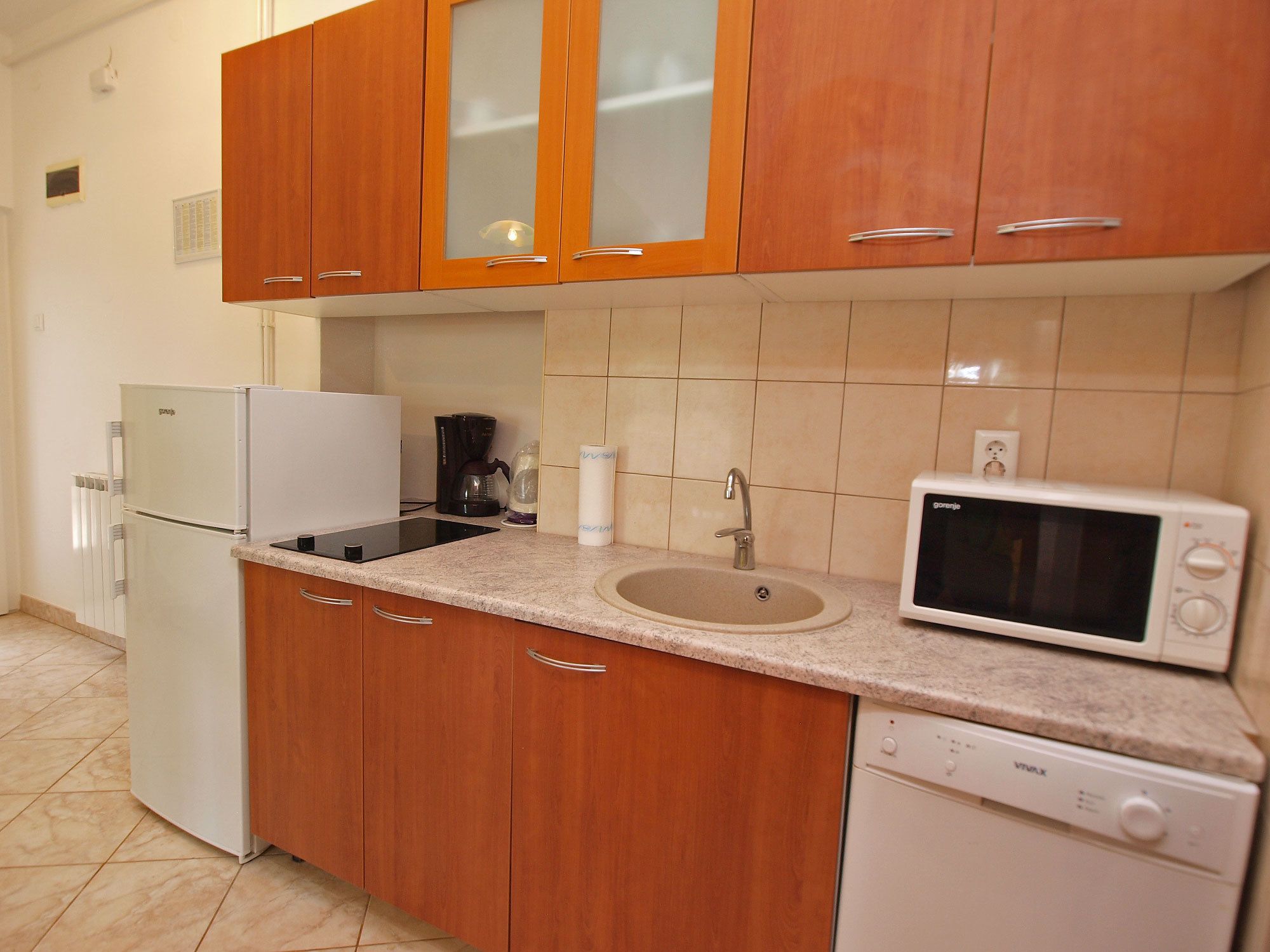 A compact kitchen with wooden cabinets, a refrigerator, a coffee maker, a microwave, and a dishwasher.
