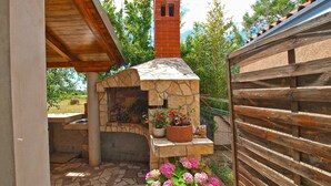 A stone-built outdoor kitchen with a chimney, surrounded by a wooden fence and potted plants. - Apartments Daffodils 1002 (Fazana)