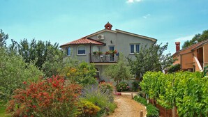 A two-story house with a red-tiled roof, surrounded by a garden with various plants and a gravel path leading to the entrance. - Apartments Daffodils 1002 (Fazana)