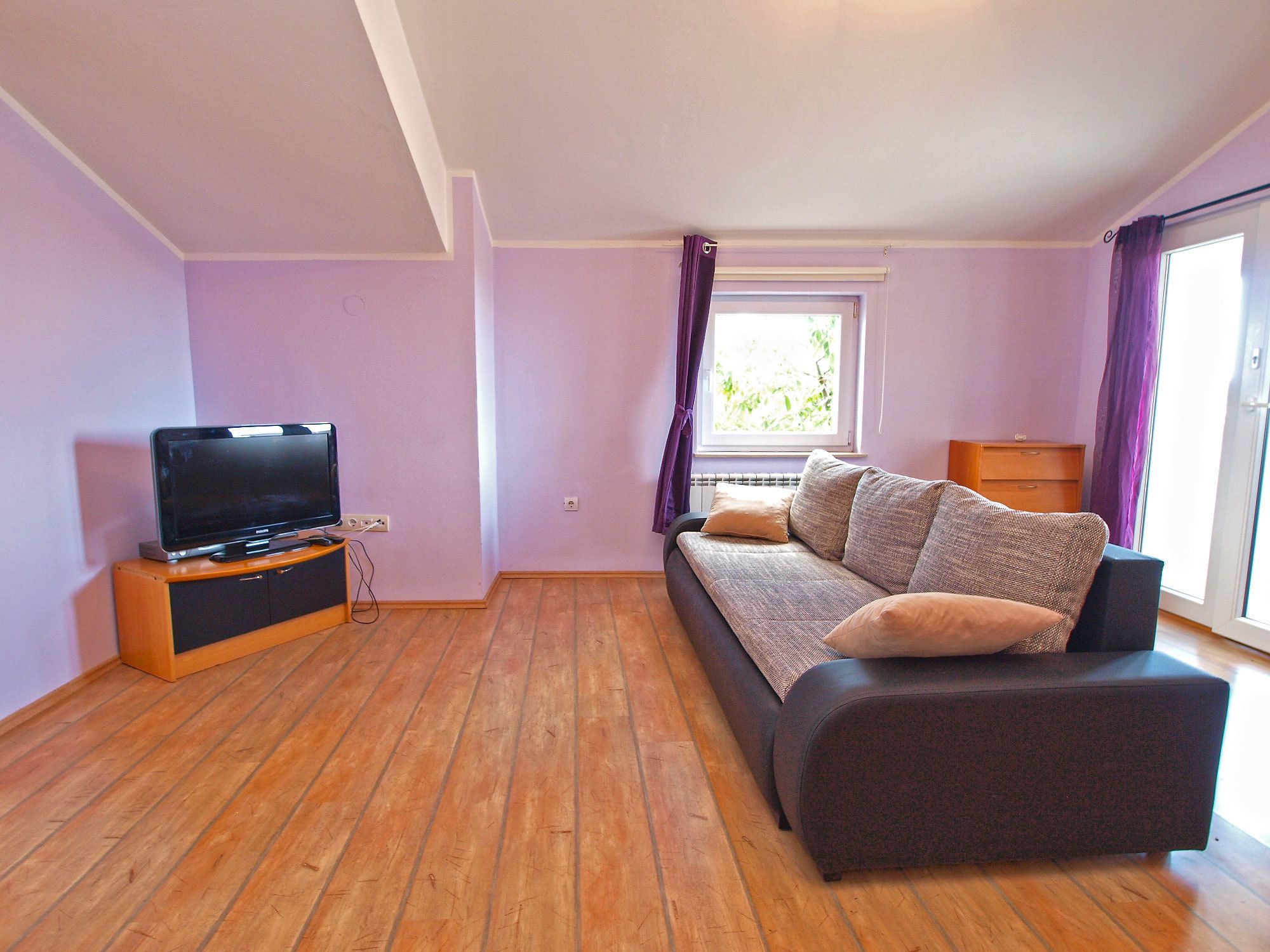 Een kamer met een bank, een televisie op een houten dressoir, een raam met gordijnen en een houten vloer.