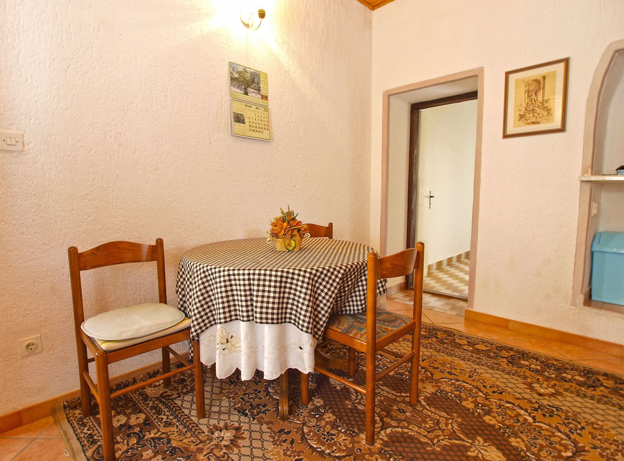 A dining area with a checkered tablecloth, two wooden chairs, a vase with flowers, and a patterned rug.