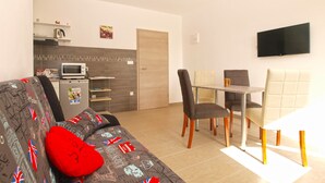 Ein kompakter Wohnraum mit Sofa, Esstisch und Stühlen. Ein Fernseher ist an der Wand befestigt, und eine Mikrowelle steht auf der Arbeitsfläche.