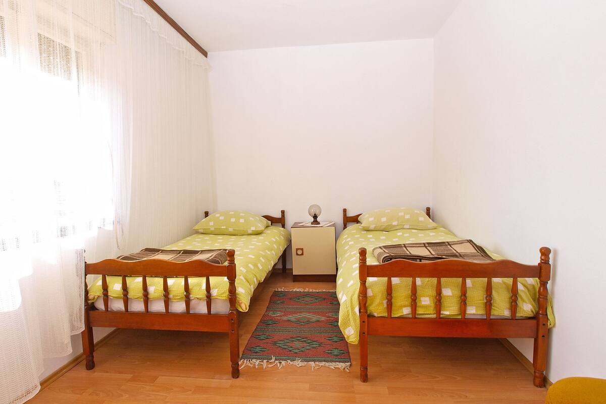 A small, clean room with two wooden beds, a nightstand, and a window with sheer curtains.