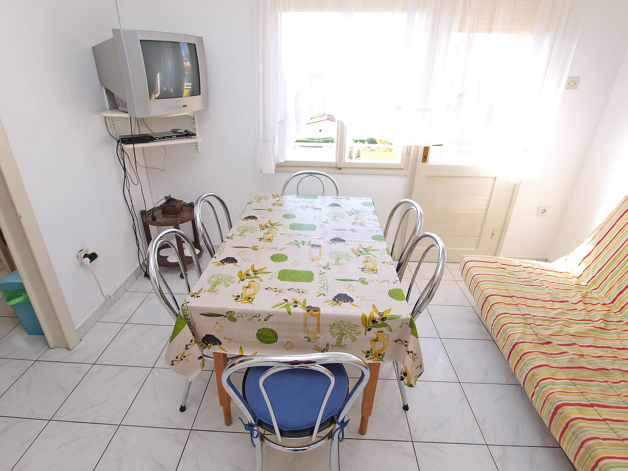A dining area with a table covered in a tablecloth, metal chairs, a television on a stand, and a window with curtains.