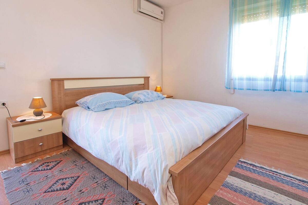 A bedroom with a wooden bed, two bedside tables with lamps, a window with sheer curtains, and a wall-mounted air conditioner.