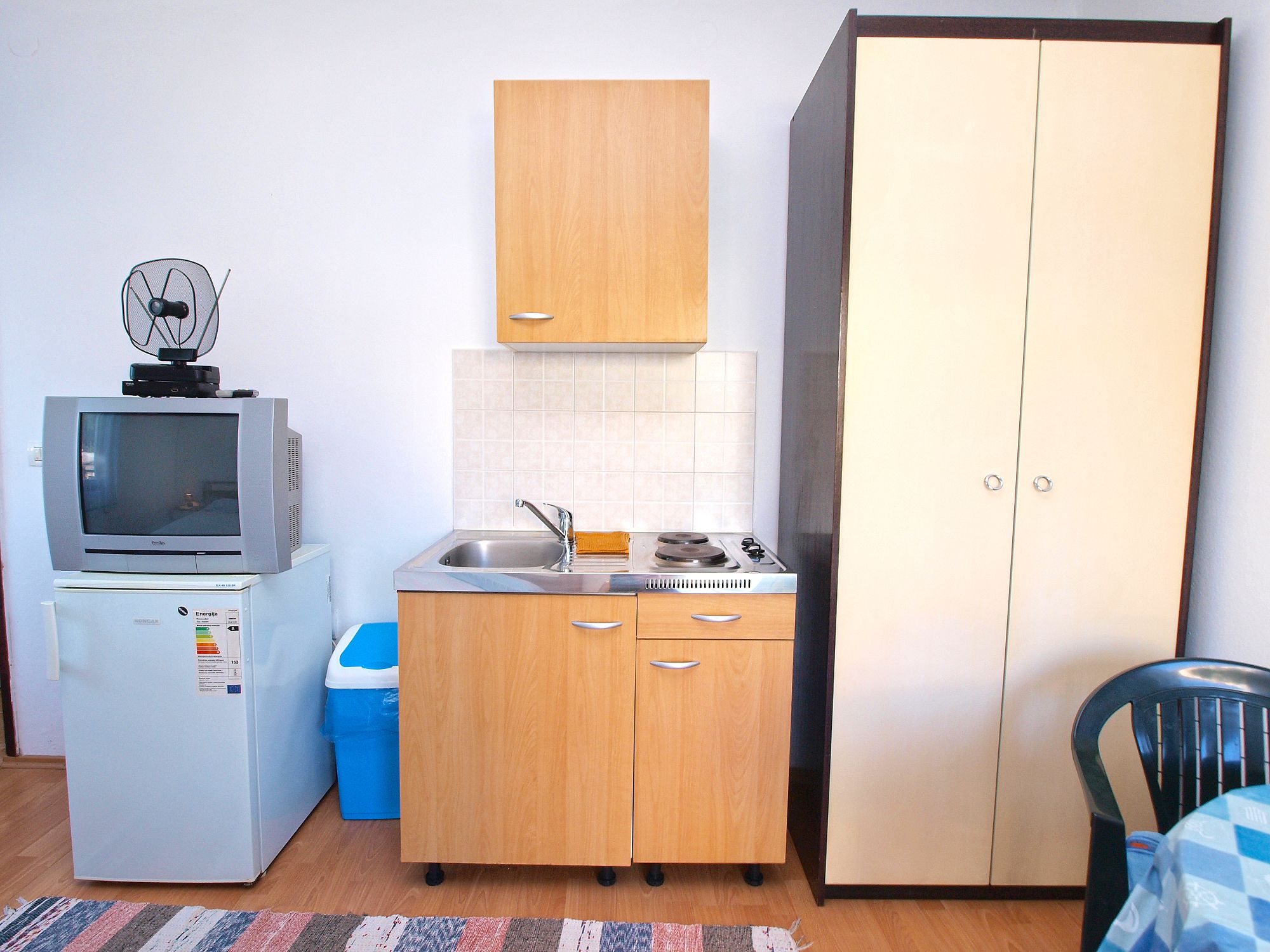 A compact kitchenette with a refrigerator, a microwave, a sink, and a stove. There is a wooden cabinet above the sink and a large wardrobe to the right.