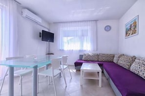 A modern living room with a glass dining table, white chairs, a purple sofa, and a mounted TV.