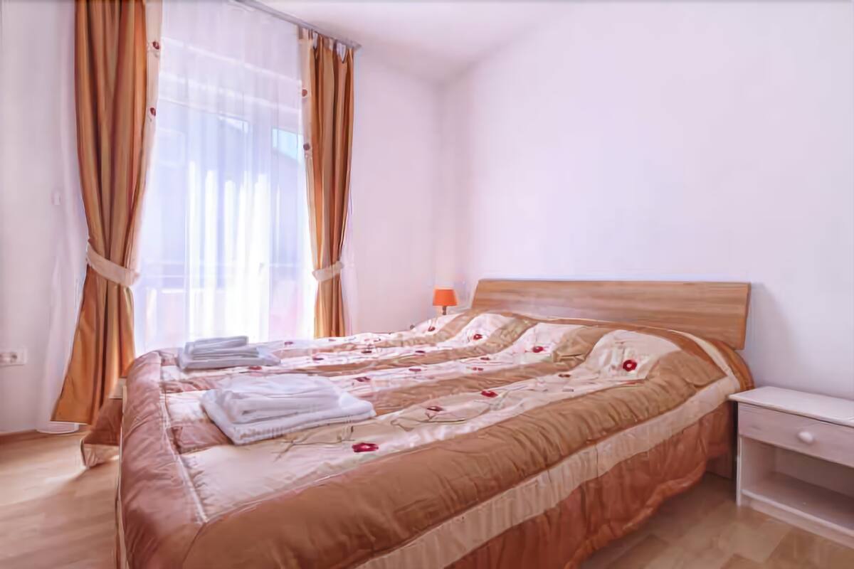 A neatly made bed with a floral duvet, a bedside table with a lamp, and sheer curtains.