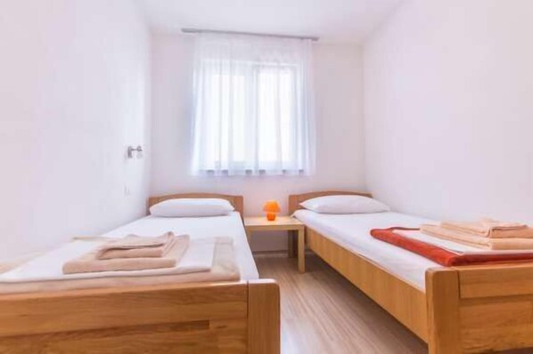 A small, clean room with two wooden beds, a nightstand with a lamp, and a window with sheer curtains.