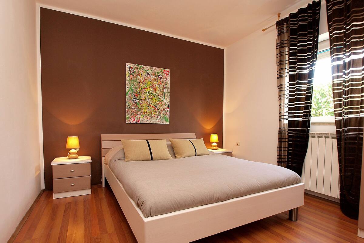 A bedroom with a bed, two bedside tables, a wall-mounted lamp, and a window with curtains.