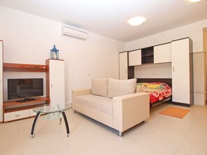 A compact living space with a sofa, a glass table, a TV, and a bed with a colorful blanket. - Apartments Elvis 1299 (Vrsar)