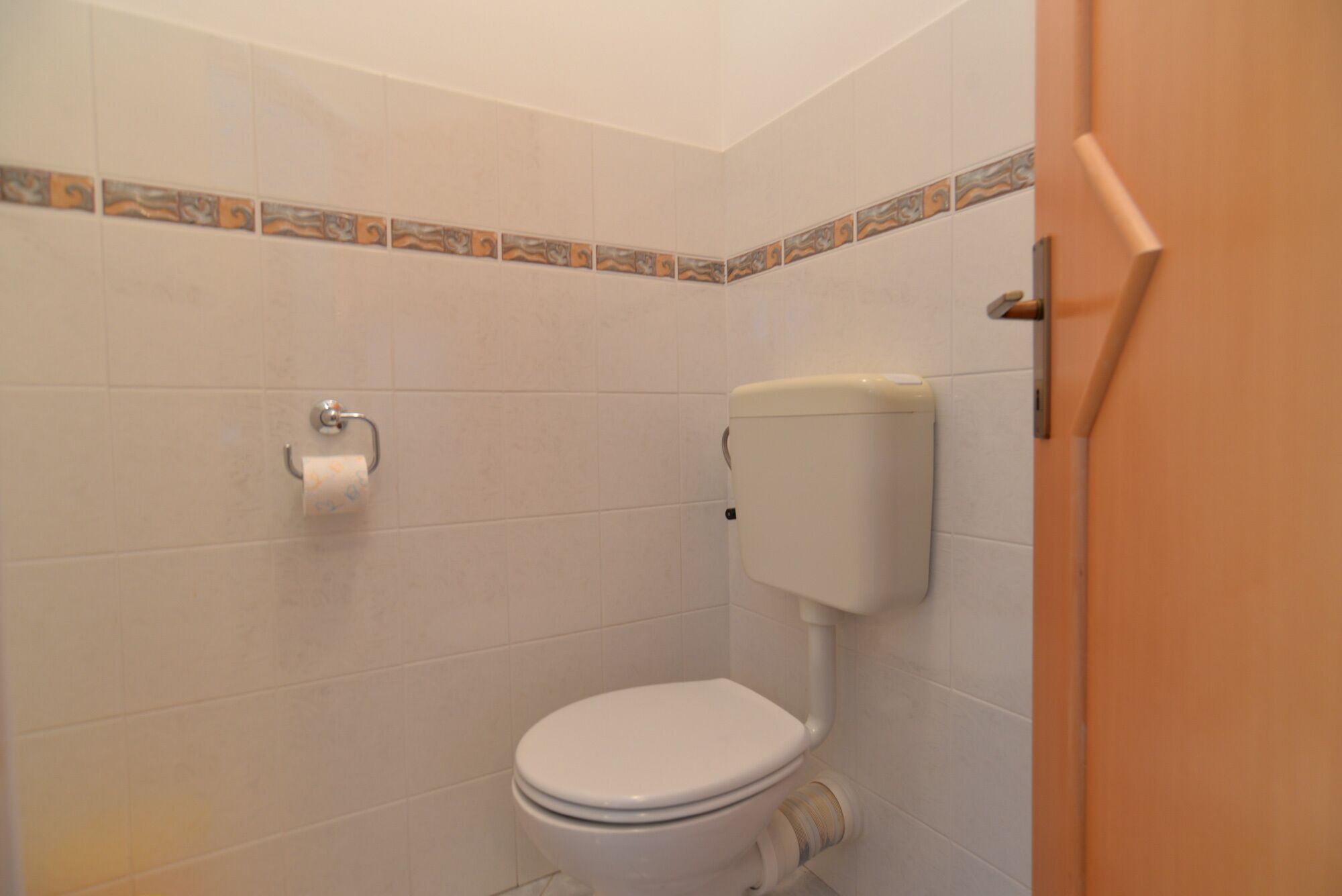 Une petite salle de bain propre, équipée de toilettes, d’un porte-rouleau de papier toilette et d’un mur carrelé.