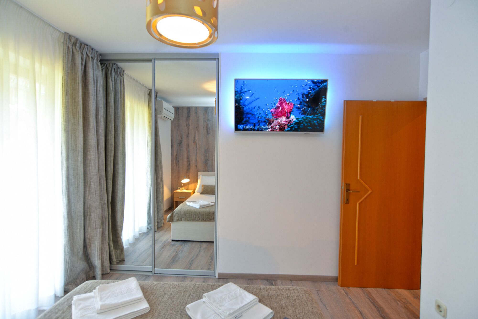A modern hotel room with a large mirror, a flat-screen TV, a wooden door, and a view of the adjacent room.
