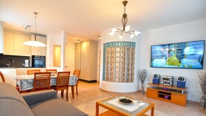 Una sala de estar moderna con sofá, mesa de centro y televisor. Incluye una zona de cocina con armarios y una mesa de comedor.