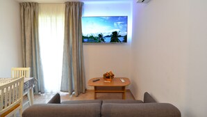 Una sala de estar con un sofá, una mesita con un florero, un televisor en la pared y una ventana con cortinas.